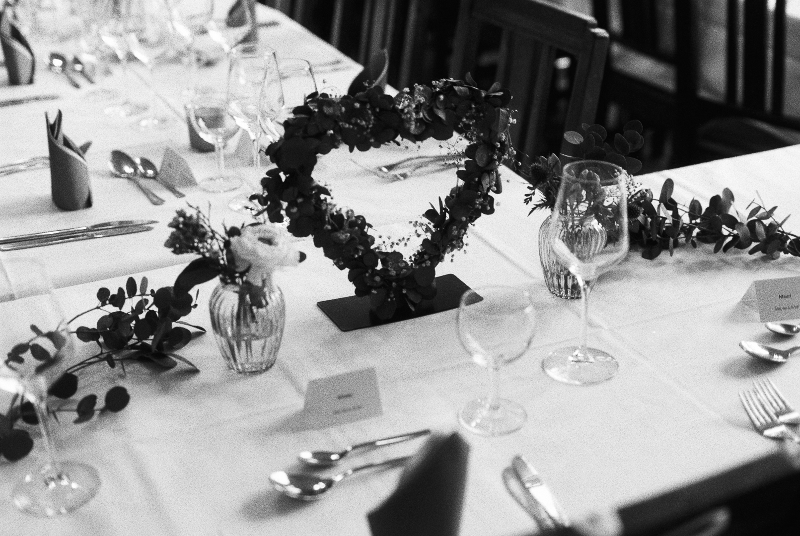 Ein elegant gedeckter Hochzeitstisch mit weißen Tischdecken, filigranen Weingläsern und stilvoll arrangierten Bestecken. In der Mitte steht ein herzförmiger Blumenkranz aus Eukalyptus und zarten Blüten, flankiert von kleinen Vasen mit frischen Blumen. Die liebevollen Details und die romantische Tischdekoration vermitteln eine festliche Atmosphäre und eignen sich perfekt für die visuelle Darstellung einer modernen, stilvollen Hochzeit. Dieses Bild zeigt genau die Art von Momenten und Details, die ein Hochzeitsfotograf Saarland professionell und emotional einfangen kann.